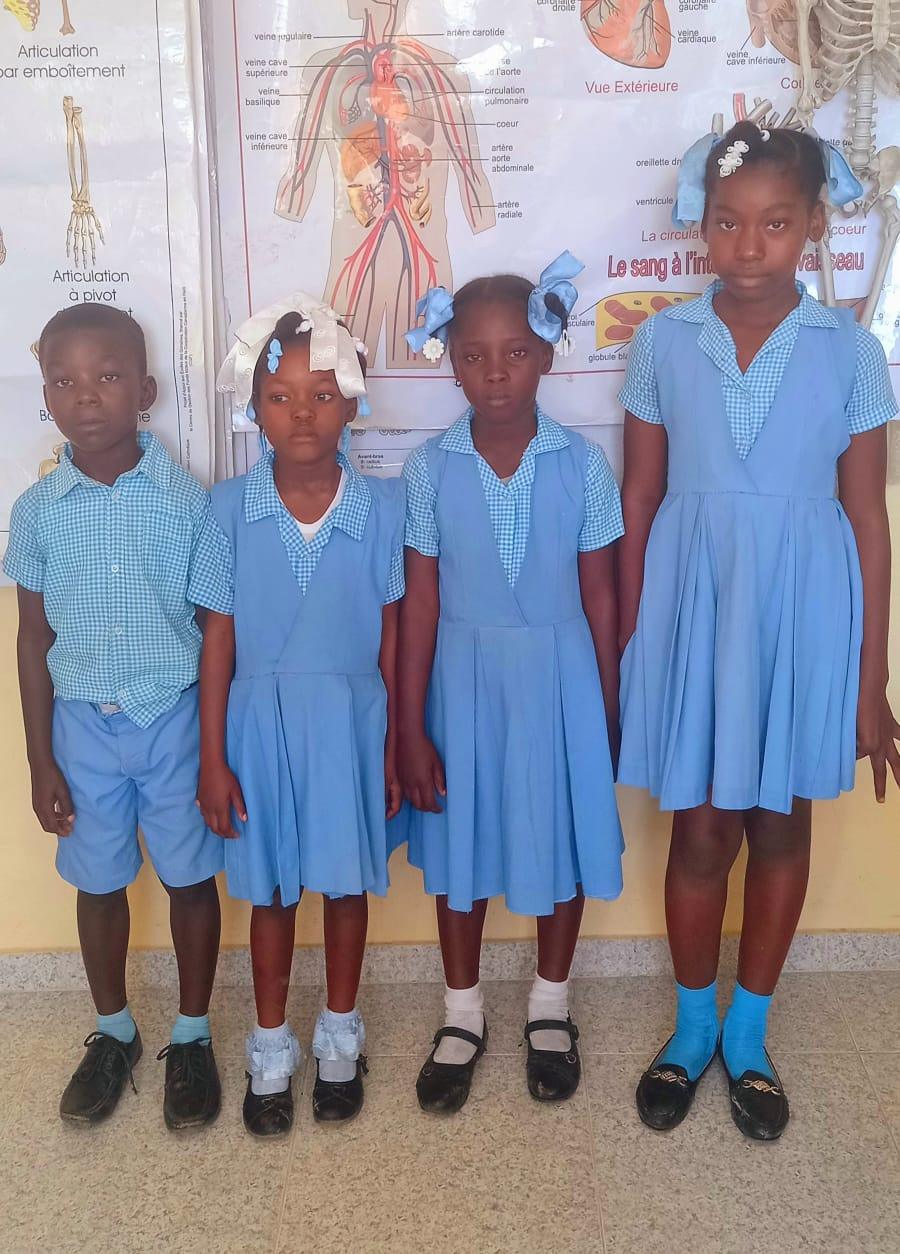 Four children in school uniform, supported by HTHH, standing in a classroom at a partner school in Haiti.