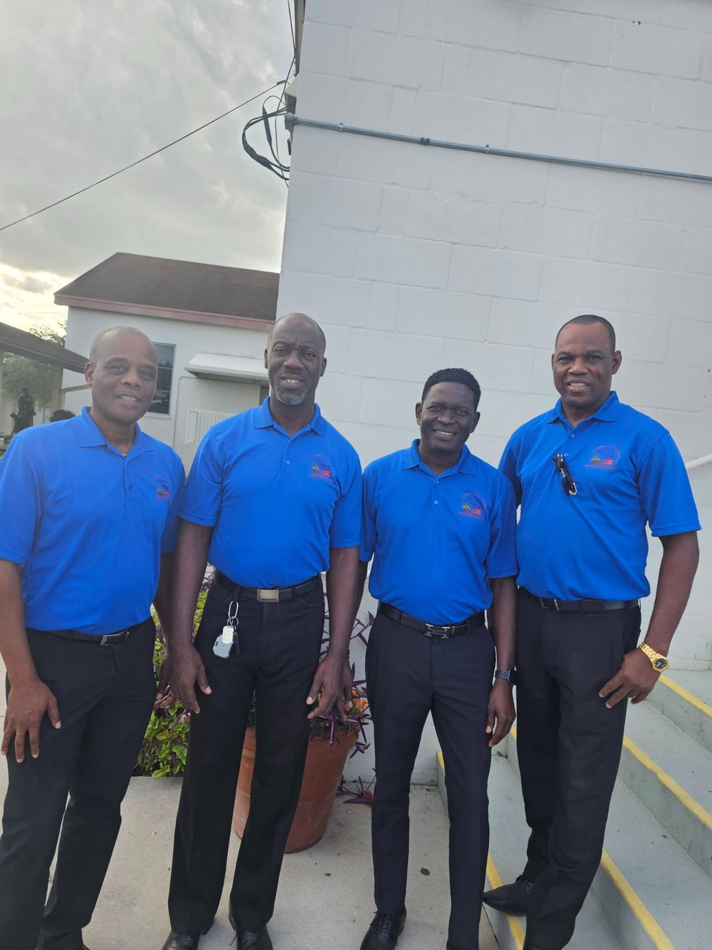 Four HTHH volunteers in blue branded shirts standing outside a community building.