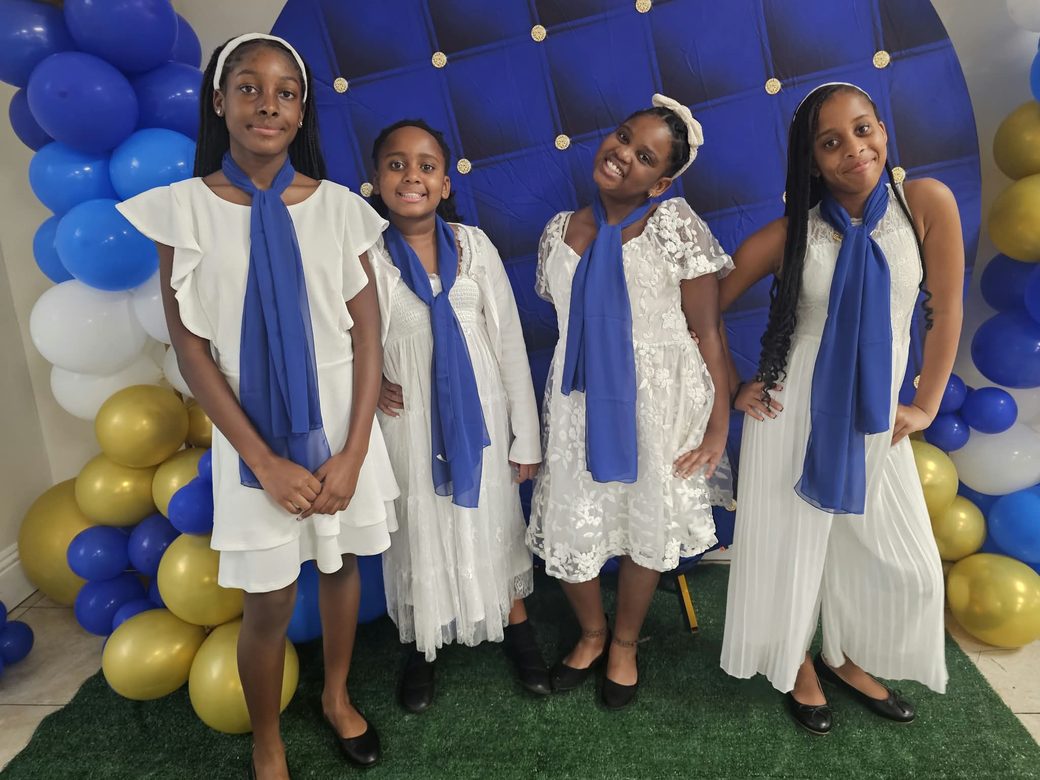 Four girls in white dresses with blue sashes at an HTHH youth event.